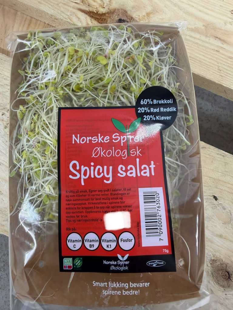 En pakke Norske Spirer Spicy salat i et avlangt pappbeger med plast rundt og rød etikett.
